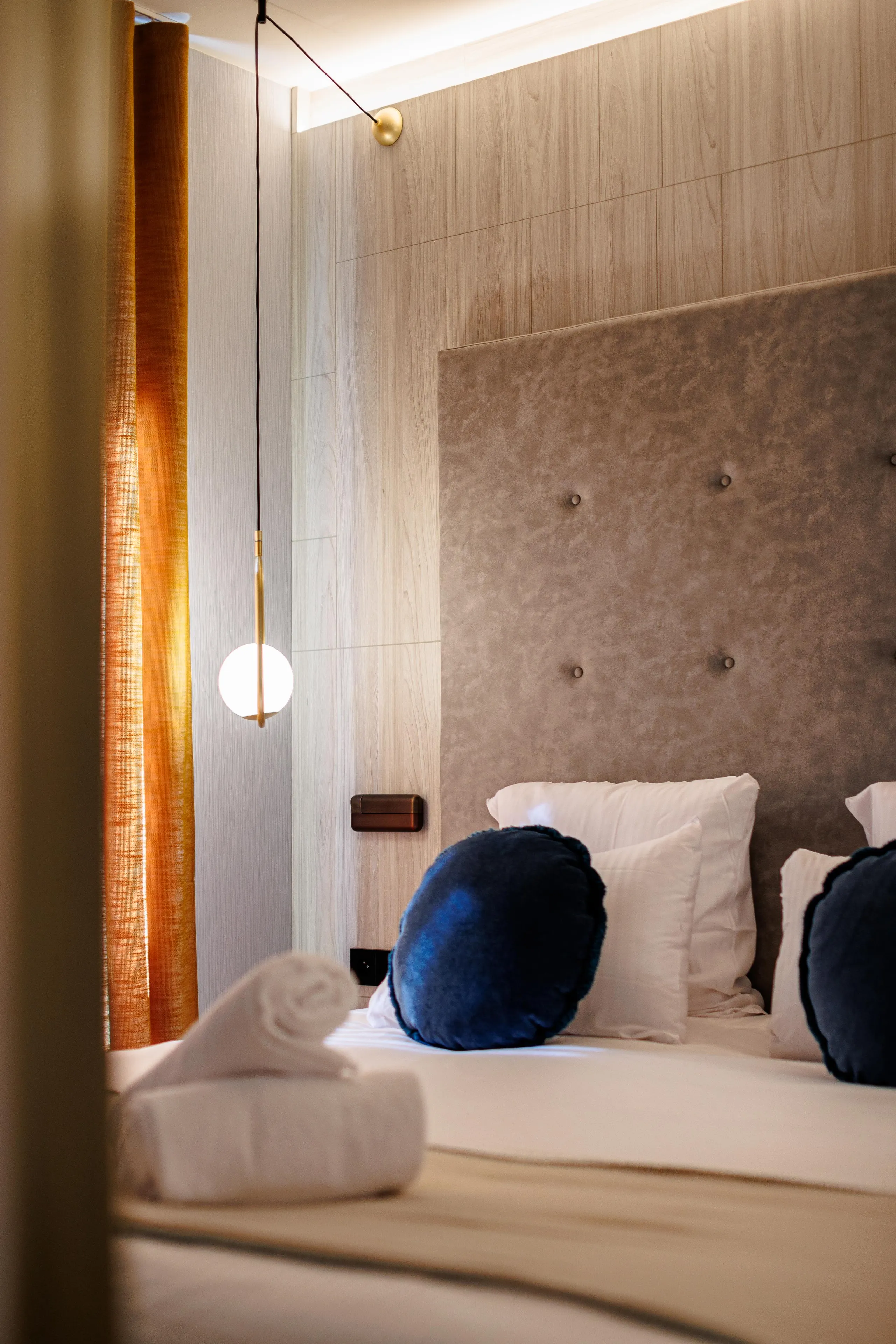 Bed with a fabric headboard and cushions, a hanging lamp, and a window with an orange curtain