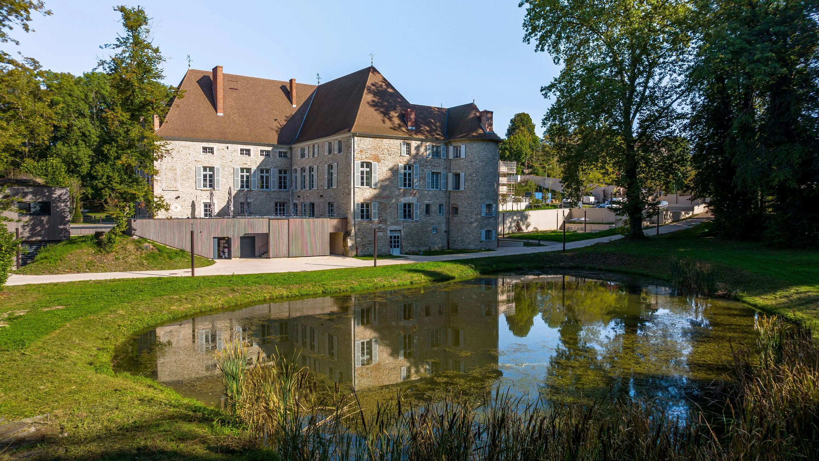 Old stone building with red-tiled roofs surrounded by trees, reflected in a pond surrounded by grass and vegetation.
