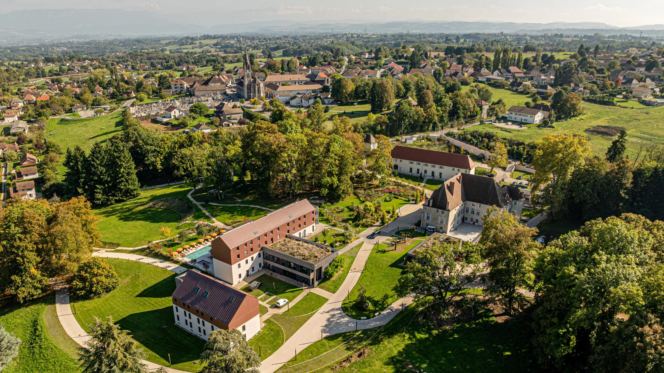 Aerial view of a group of buildings surrounded by greenery in a village with fields and houses in the background