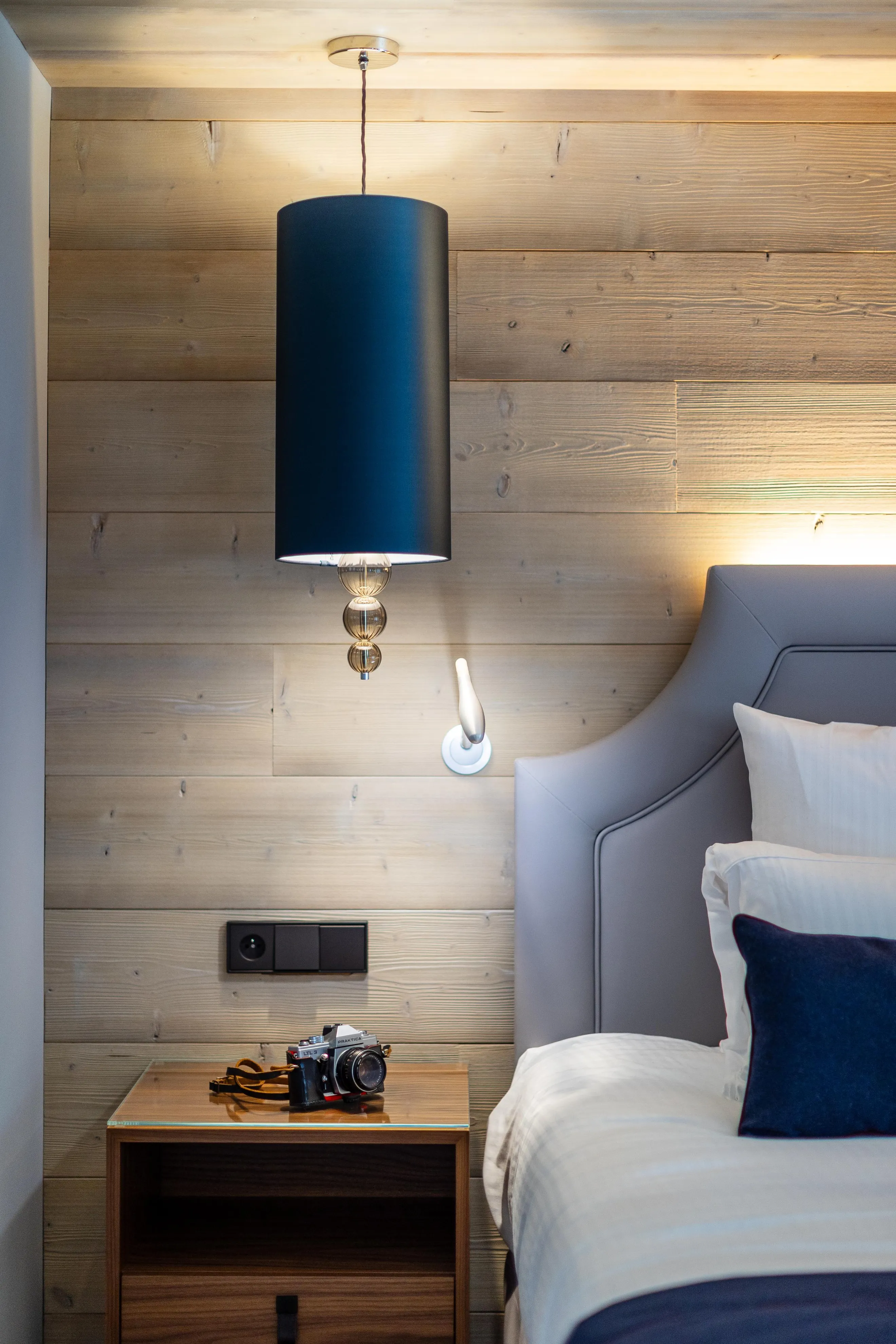A room with a bed, a hanging lamp, a bedside table with a camera and a wall lamp.