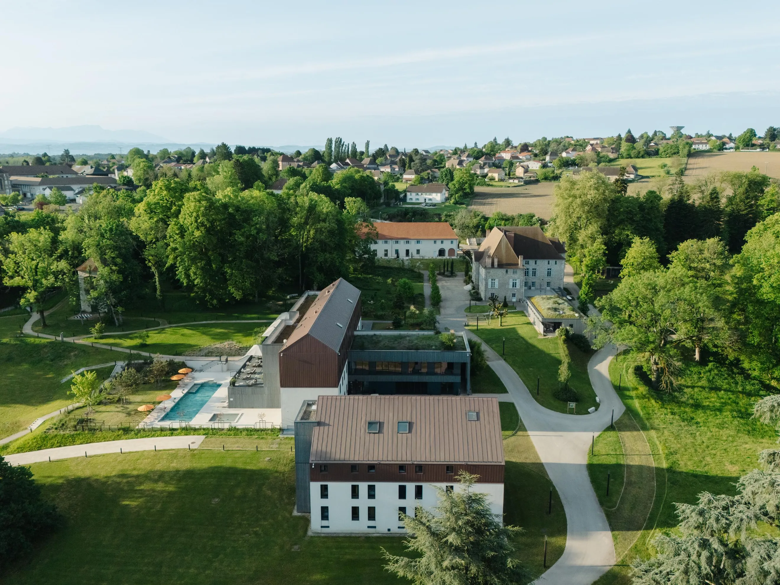 Aerial view of a domain with buildings, pools, and gardens surrounded by trees and paths, on the edge of a village and fields.