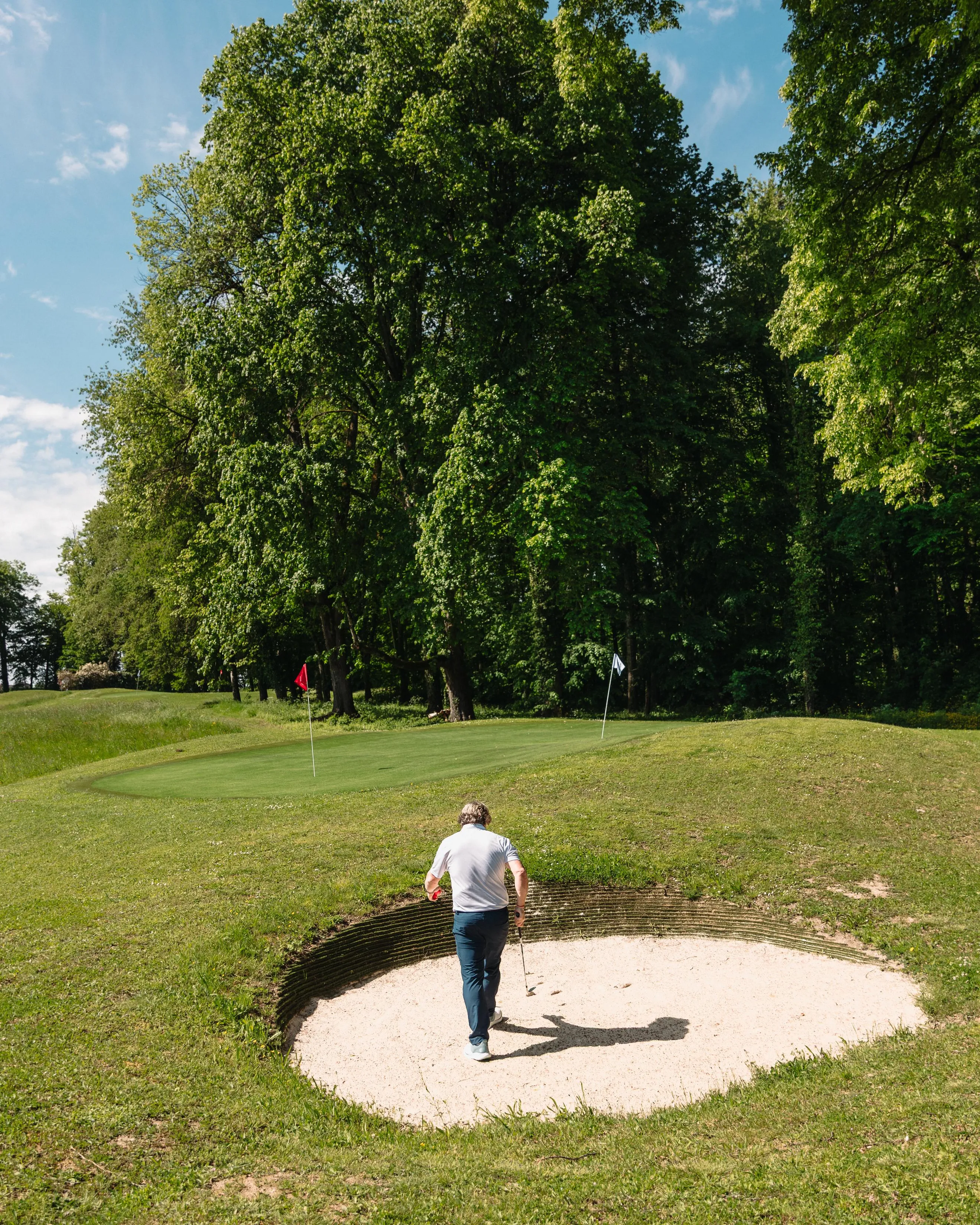 A golfer stands on a sand bunker and looks toward the green surrounded by trees.