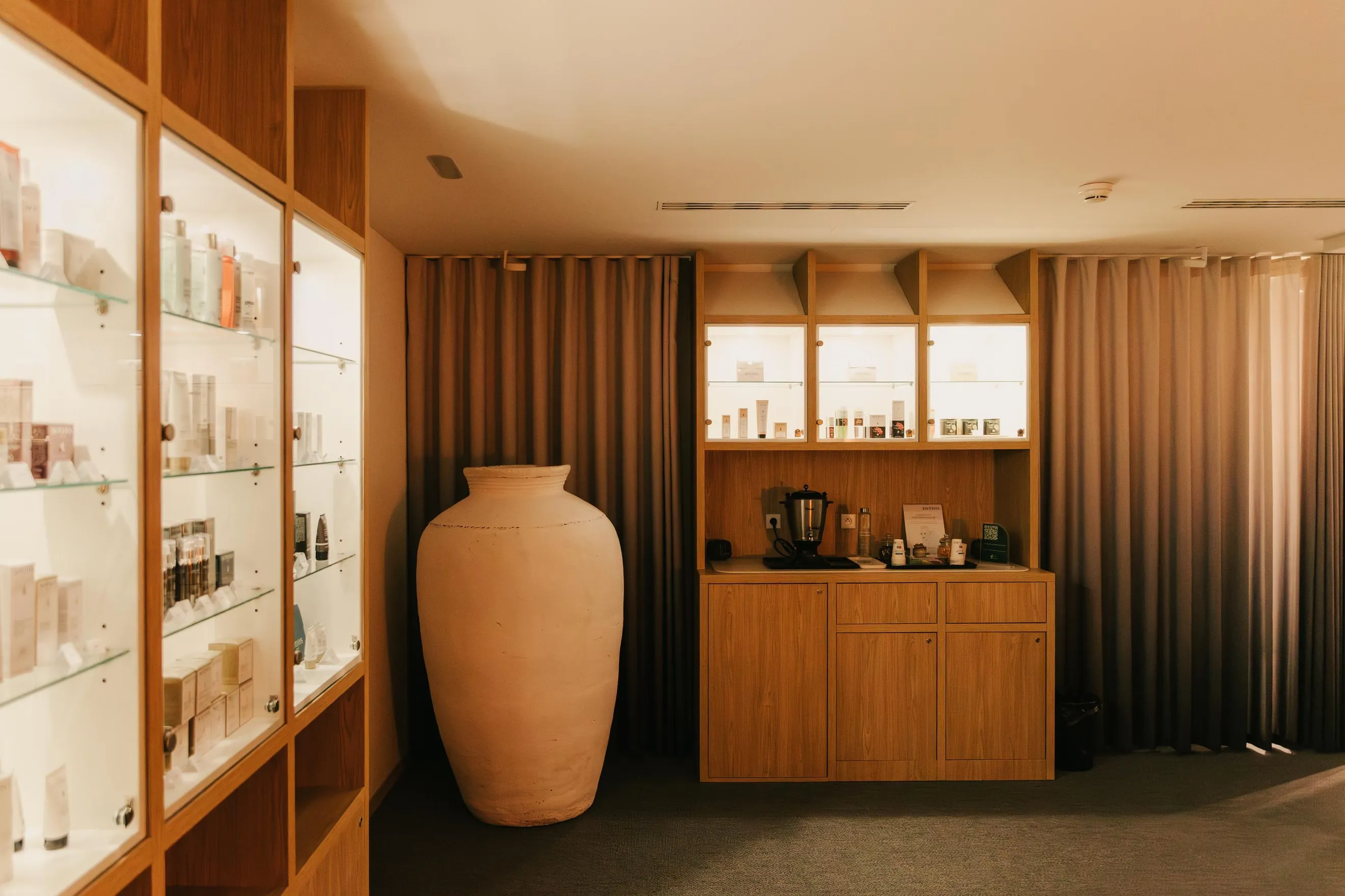 A large terracotta vase is placed in front of a wooden piece of furniture with glass shelves filled with products and a counter with various objects.