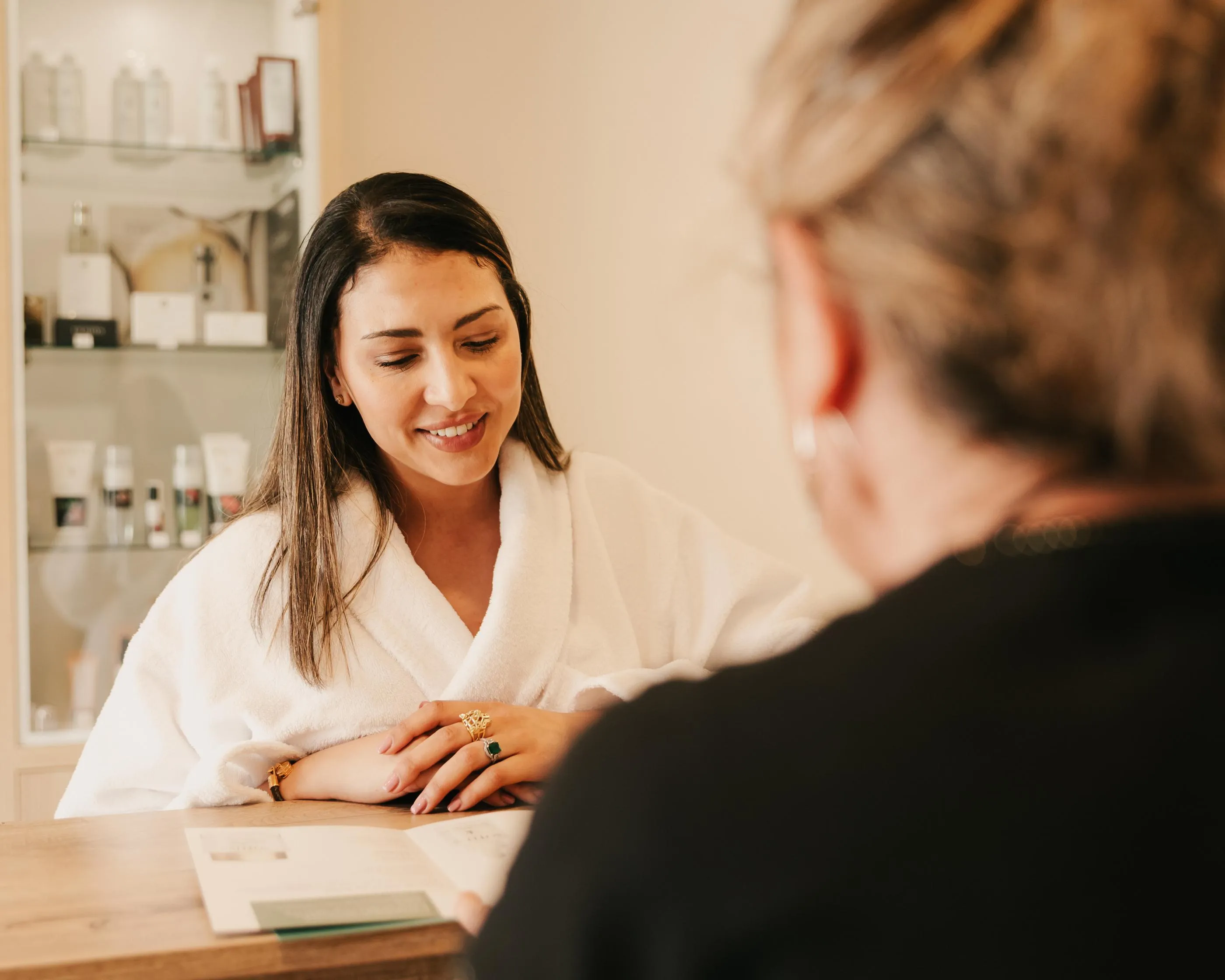 A woman in a white swimsuit smiles and talks to a person whose back is visible, in a beauty salon with products in the background.