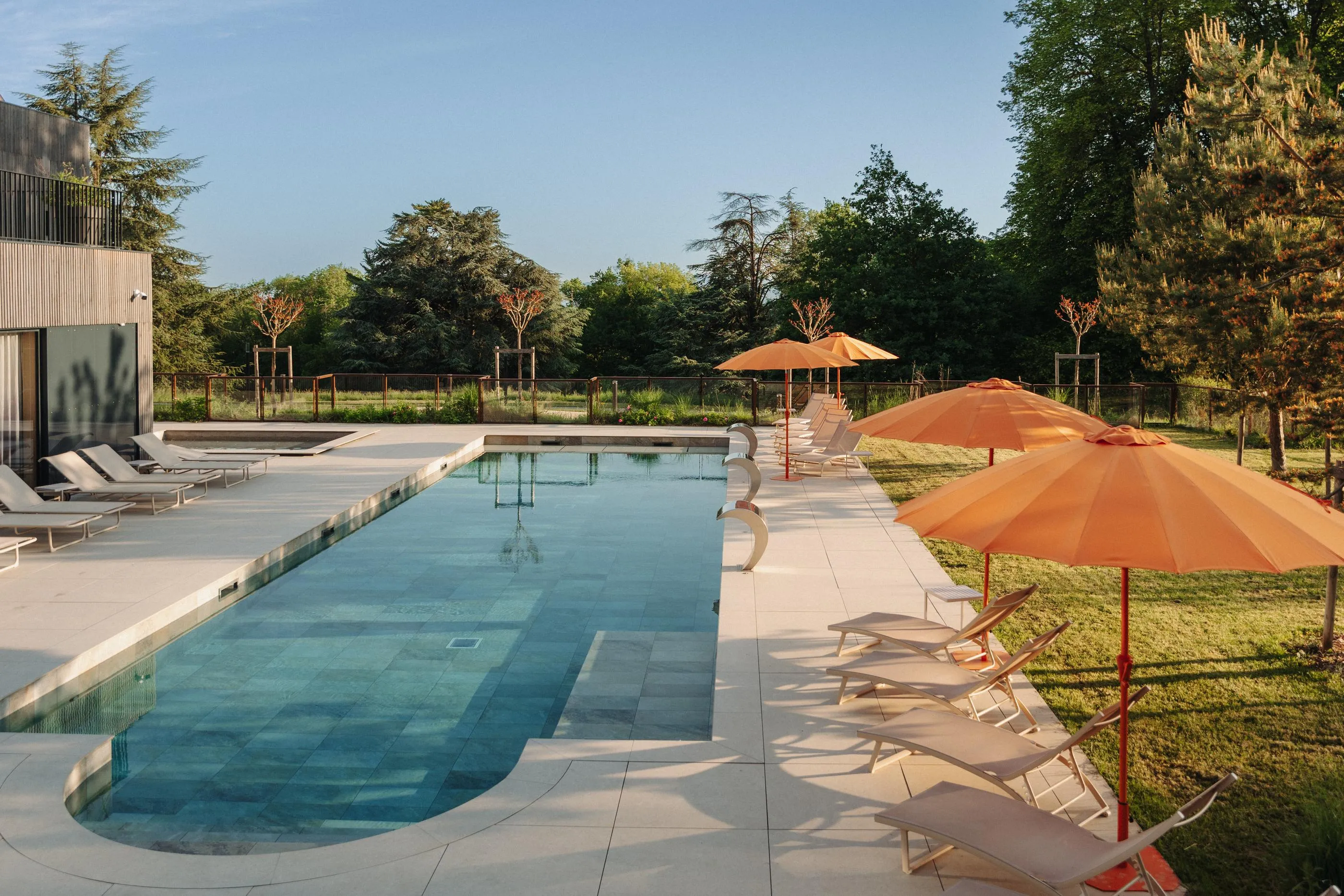 Rectangular swimming pool surrounded by sun loungers and parasols under a clear sky with trees in the background